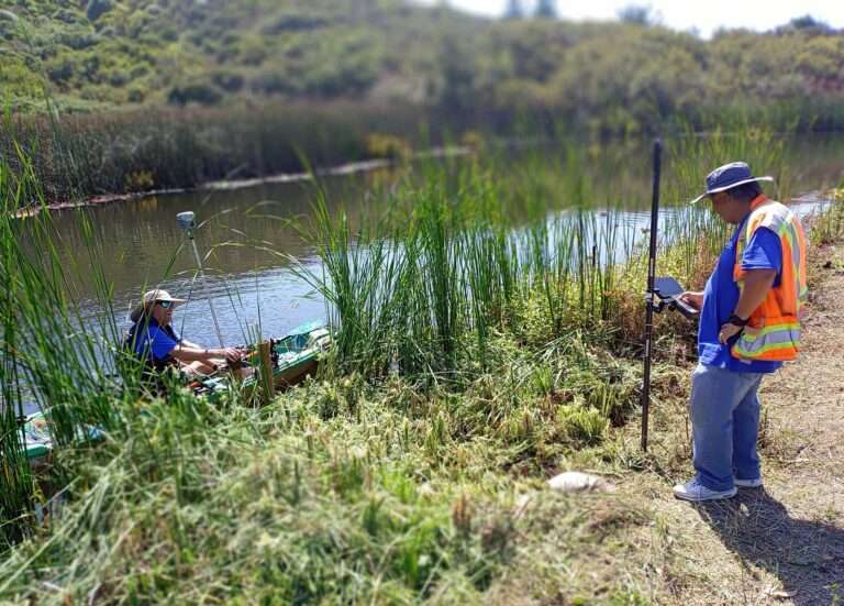 Two surveyors in orange vests and holding gear. One standing on the bank of the water and one in a kayak in the water.